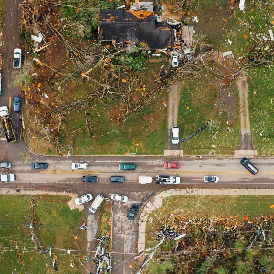 An overhead view of tornado damage along a street: a roof torn open, snapped trees and debris scattered across gardens and the road.